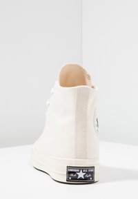 Høytopp kanvas sneakers i off-white med teksturert overflate, en svart merkepatch på hælen, og sølv øyer til snøring.