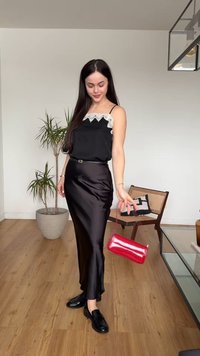 Femme en jupe et haut en satin noir avec bordure en dentelle, tenant un sac à main rouge, debout sur un sol en bois près d'une plante et d'une chaise avec coussin.