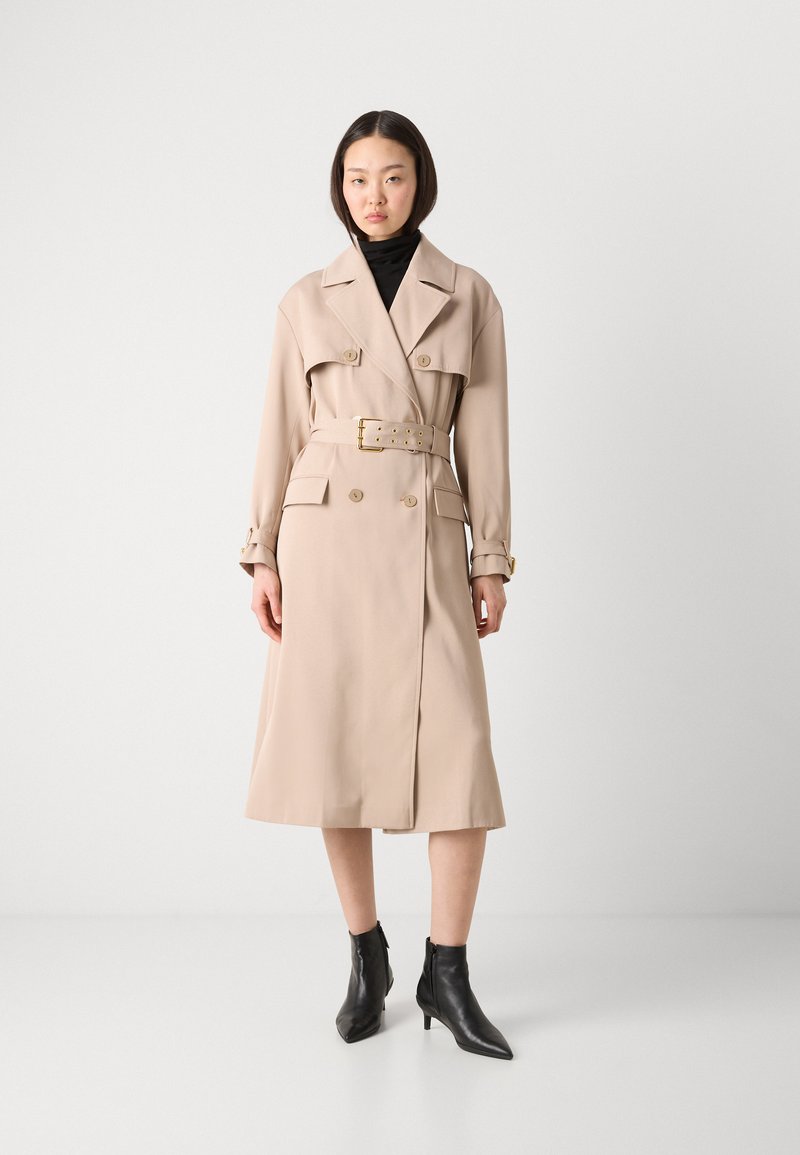 Beige Trenchcoat mit doppelt geknöpften Knöpfen, tailliertem Gürtel und zwei vorderen Taschen. Getragen über einem schwarzen Rollkragenpullover und kombiniert mit schwarzen Ankle Boots.