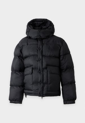 Jeune homme portant une doudoune noire, un pull beige, un pantalon large foncé et des chaussures noires, debout devant un fond clair uni.