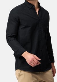 Chemise noire boutonnée avec manches longues, col et poche poitrine gauche. Le tissu semble lisse avec une texture mate.