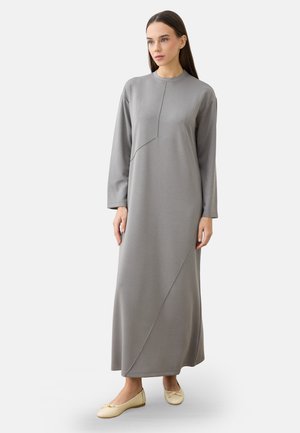 Woman with long brown hair wearing a long-sleeve, ankle-length gray dress and beige ballet flats standing against a plain background.