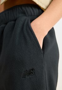 Melnas flīsa bikses ar elastīgu jostu un sānu kabatām; kreisajā augšstilbā ir izcilts New Balance logo.