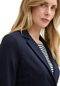 Blazer marin cu rever structurat, design ajustat și textură netedă; purtat peste o cămașă cu dungi. Liniile moi, elegante pun în evidență silueta.