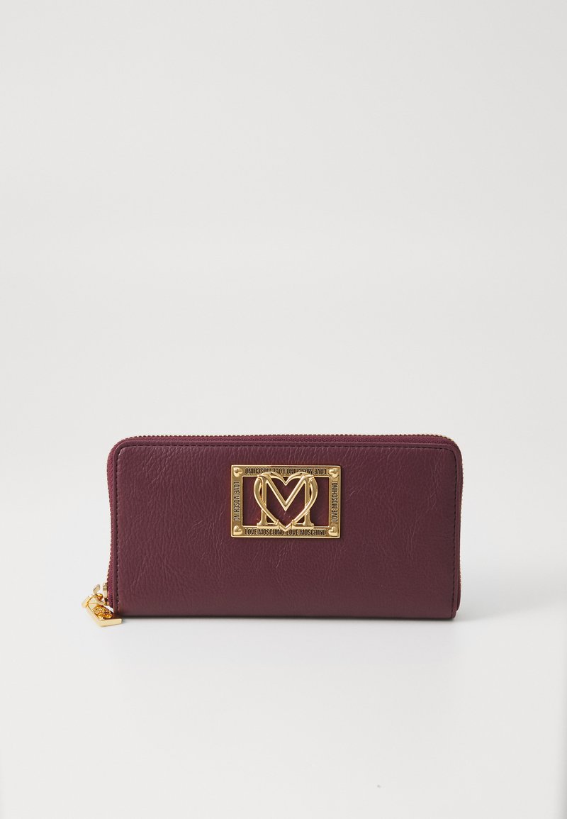 Burgundy textured leather wallet with a gold-tone logo plaque, zip closure, and a small charm at the zipper pull.