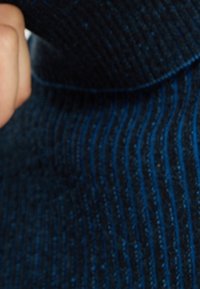Primo piano di una mano che sistema il colletto di un maglione di lana a coste blu scuro con righe verticali.
