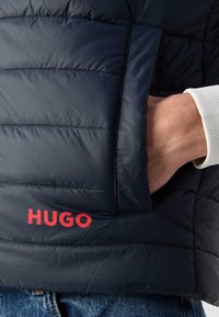 Granatowa kurtka puchowa z pikowanym wzorem, z wyraźnym czerwonym logo "HUGO" na dolnym lewym boku oraz widoczną boczną kieszenią.