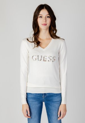 Maglione bianco a maniche lunghe con scollo a V e logo "GUESS" in strass luccicanti sul petto, abbinato a jeans blu.