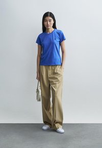 Sinine lühikeste varrukatega puuvillane T-särk, kombineeritud beežide voldiliste laiade pükstega ja valgete tennistega, hoides väikest valget kotti.