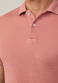 Polo tričko v jemnej ružovej farbe s textúrovaným úpletom. Má preložený golier, dva gombíky pri krku a krátke rukávy.