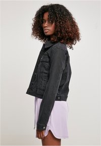 Zwarte denimjack met een cropped ontwerp, voorzakken en drukknoopsluiting. Gestyled over een lichtpaarse jurk met een relaxte pasvorm.