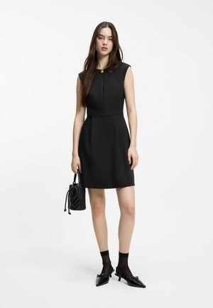 Ung kvinde i ærmeløs sort kjole, der holder en sort quiltet håndtaske, iført sorte sokker og spidse sko med lav hæl på hvid baggrund.