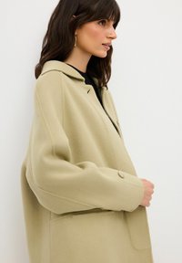 Cappotto di lana verde chiaro con una texture morbida, dotato di colletto, chiusura a un bottone e tasche grandi. Modello mostrato di profilo.