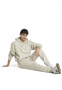 Felpa beige con tasca frontale e logo, abbinata a pantaloni jogger coordinati. Tessuto morbido, vestibilità comoda e dettagli di design minimalisti.