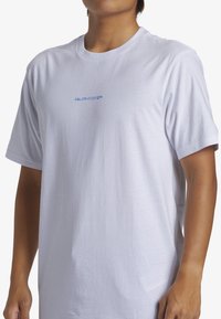 Camiseta de algodón blanca con mangas cortas, que presenta un logo azul "QUIKSILVER" en el pecho. Diseño simple y clásico con un corte holgado.