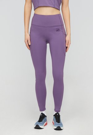 Person iført høj taljede lilla leggings med sidelommer og blå, røde og sorte sportssko mod en ensfarvet baggrund.
