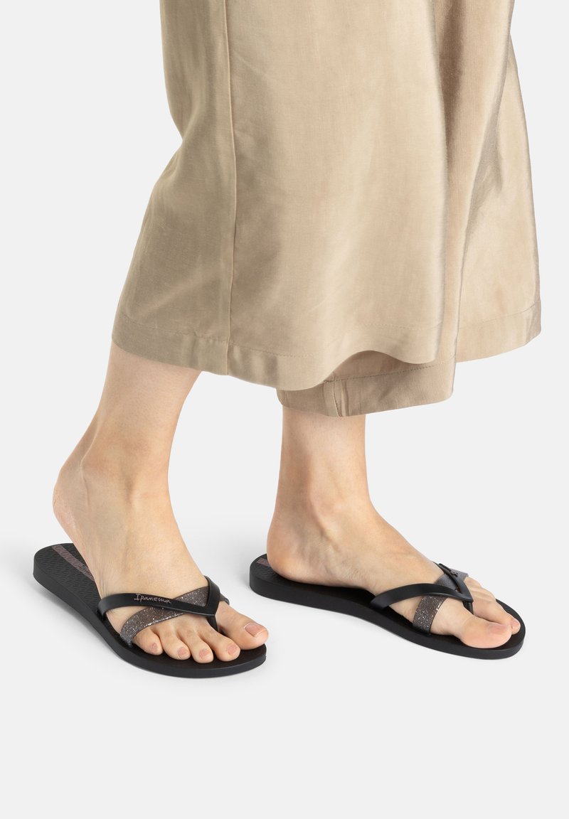 Schwarze Flip-Flops mit einer strukturierten Sohle und funkelnden Riemen, die ein minimalistisches Design aufweisen. Die Riemen kreuzen sich in Form eines X über dem Fuß.