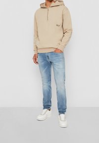 Beige mikina na zip s kapucí, se vzorem loga. V kombinaci se světle modrými džíny. Model má na sobě bílé tenisky s šedými detaily.