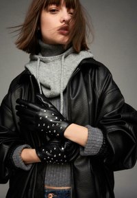 Črne usnjene rokavice s srebrnimi zakovicami, nošene na modelu s sivim puloverjem in črno usnjeno jakno, poudarjene na nevtralni podlagi.