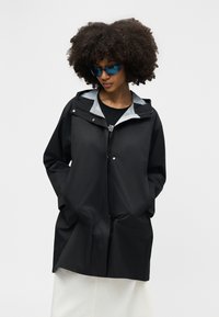 Sieviete ar cirtainiem matiem, valkā zilu saulesbrilles, melnu kapuci ar kabatās ieliktiem rokām un baltu svārku, uz vienkrāsaina fona.