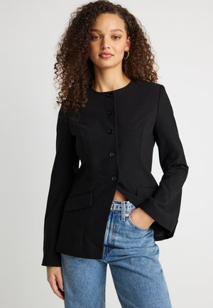 Noor naine lokkis juustega kannab kinnitatud musta bleiserit ja helesiniseid teksaseid, ühe käega taskus, valgel taustal.