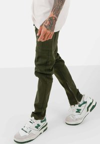 Olijfgroene cargobroek met tapered fit, voorzien van zijzakken en ritsen bij de enkels, gecombineerd met wit-groene sneakers.