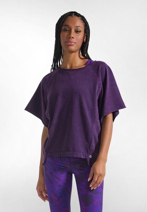 Kvinde med flettet hår iført en løs lilla top og mønstrede lilla leggings, der står mod en ensfarvet lys baggrund.