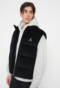 Čierna kordurová puffová vesta so zipsovým zapínaním, s bielym logom Jumpman. Nosená cez šedú mikinu, zdôrazňujúca vrstvený vzhľad.