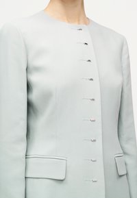 Φωτεινό γκρι σακάκι ραμμένο καλαίσθητα με μακριά μανίκια, χωρίς γιακά, με επτά κλεισίματα μπροστά με γάντζο και ματάκι, που διαθέτει δύο τσέπες με καπάκι.