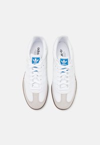 Vita sneakers med en slät läderöverkonstruktion, ljusgrå mockadetaljer, blå logotagg och brun gummisula med ett strukturerat mönster.