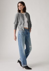 Grijze wollen cardigan over een witte slim-fit shirt, gecombineerd met lichtblauwe versleten jeans en zwarte rechte enkellaarsjes. Model poseert in een neutrale setting.