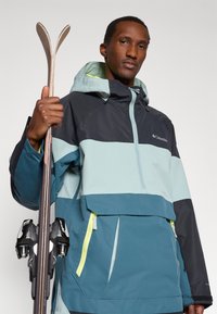 Jaqueta de esqui leve em tons de azul com detalhes em preto, apresenta capuz, fecho frontal e zíperes em amarelo néon. Segura os esquis com uma mão.