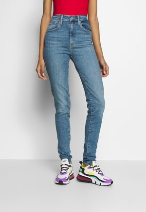 Jeans bleus taille haute en denim, avec une coupe slim et un design à cinq poches, associés à des baskets colorées avec un motif ondulé.