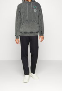 Sudadera gris de gran tamaño con bolsillo frontal, capucha con cordón y detalles laterales, combinada con pantalones rectos negros y zapatillas blancas.