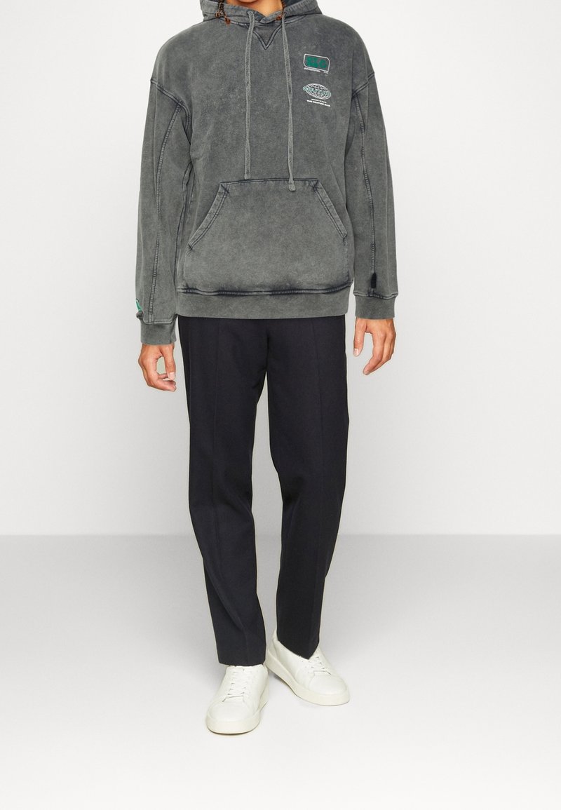 Sudadera gris de gran tamaño con bolsillo frontal, capucha con cordón y detalles laterales, combinada con pantalones rectos negros y zapatillas blancas.