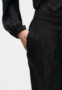 Μαύρο παντελόνι με σχέδιο, που διαθέτει λεπτή υφή, ομαλή επιφάνεια και ελαστικά τελειώματα στους αστραγάλους. Το σχέδιο περιλαμβάνει επαναλαμβανόμενο μοτίβο λογότυπου.