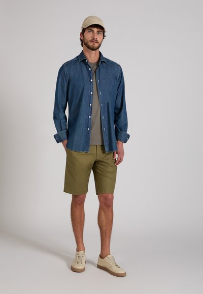 Hombre con gorra beige, camisa vaquera azul sobre camiseta gris, pantalones cortos verde oliva y zapatillas beige de pie contra un fondo liso.