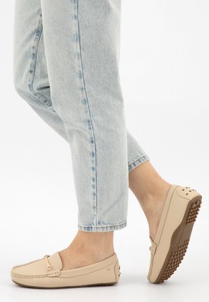 Piedi in jeans azzurri chiaro che indossano mocassini in pelle beige con suole in gomma chiodate su sfondo bianco.