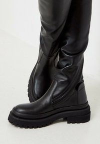 Black tall boots made of leather with a smooth texture, featuring a chunky sole, side zipper, and minimal seam detailing.