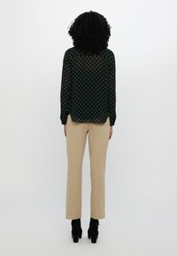 Femme aux cheveux bouclés, de dos, portant un chemisier noir à manches longues avec des pois verts, un pantalon beige et des bottes noires à talons.