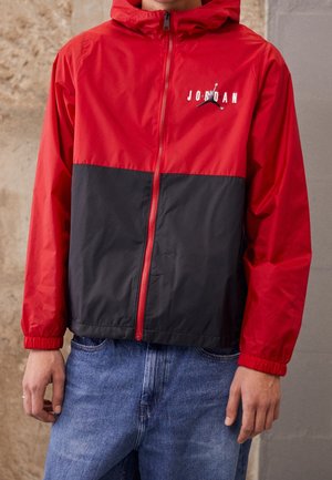 Man draagt een rood-zwart windjack met rits en capuchon, voorzien van het Jordan-logo, gecombineerd met een blauwe jeans, staand tegen een effen muur.