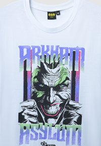 Biała bawełniana koszulka z graficznym nadrukiem twarzy Jokera z zieloną skórą, purpurowymi cieniami oraz tekstem "ARKHAM ASYLUM" w wyrazistych kolorach.