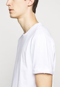 White cotton t-shirt with a crew neck, short rolled sleeves, and a relaxed fit, displayed on the side of a model against a plain background.