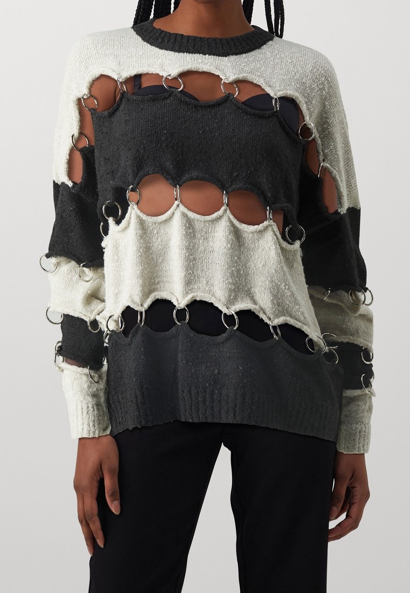 Pull tricoté en noir, gris et blanc avec des sections découpées ; comprend des accents en anneaux métalliques. Poignets côtelés et col rond.