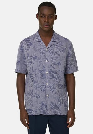 Mann trägt hellblaues Kurzarmhemd mit dunkelblauen tropischen Blattmustern und dunkle Hose, steht vor einfarbigem Hintergrund.