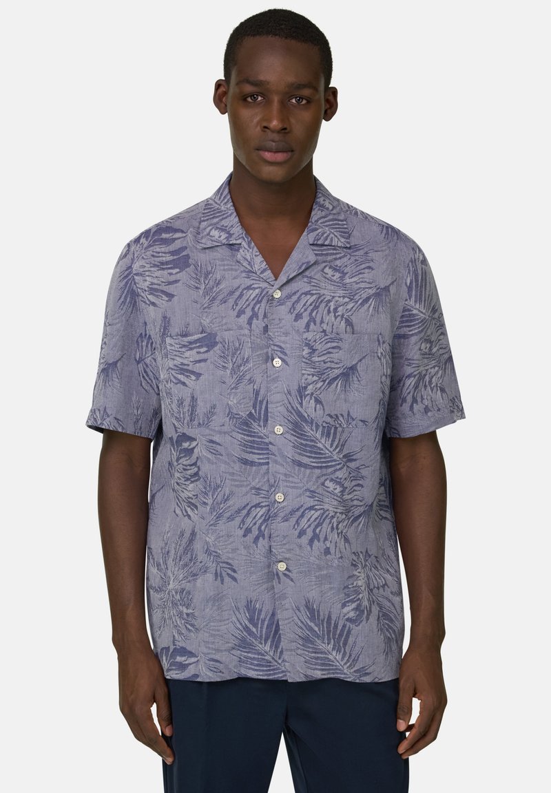 Homme portant une chemise bleu clair à manches courtes avec des motifs de feuilles tropicales bleu foncé et un pantalon foncé, debout devant un fond uni.