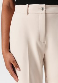 Beige tailored pants with a smooth texture, featuring a waistband, front pocket, and a silver button closure. Close-up view showing detail.