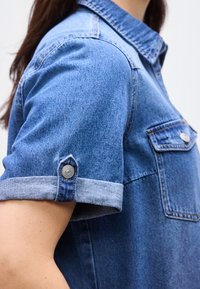 Κοντινό πλάνο ενός ατόμου που φοράει μπλε πουκάμισο από τζιν με μανίκι που έχει διπλωθεί και στερεωθεί με κουμπί και τσέπη στο στήθος.