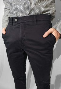 Pantaloni chinos in misto cotone nero con una texture liscia. Presentano tasche frontali, chiusura con bottone e dettagli di cuciture sottili. Indossati con una camicia a fantasia.