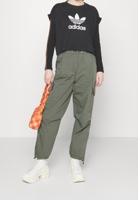 Person wearing black Adidas cropped top, olive green cargo pants, white chunky sneakers, and holding an orange plaid handbag by their side.